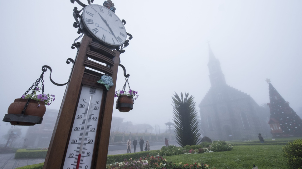 Relógio com termômetro em primeiro plano e Igreja São Pedro parcialmente encoberta pela neblina em Gramado.